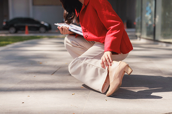 Woman experiencing ankle pain outdoors while holding documents 
